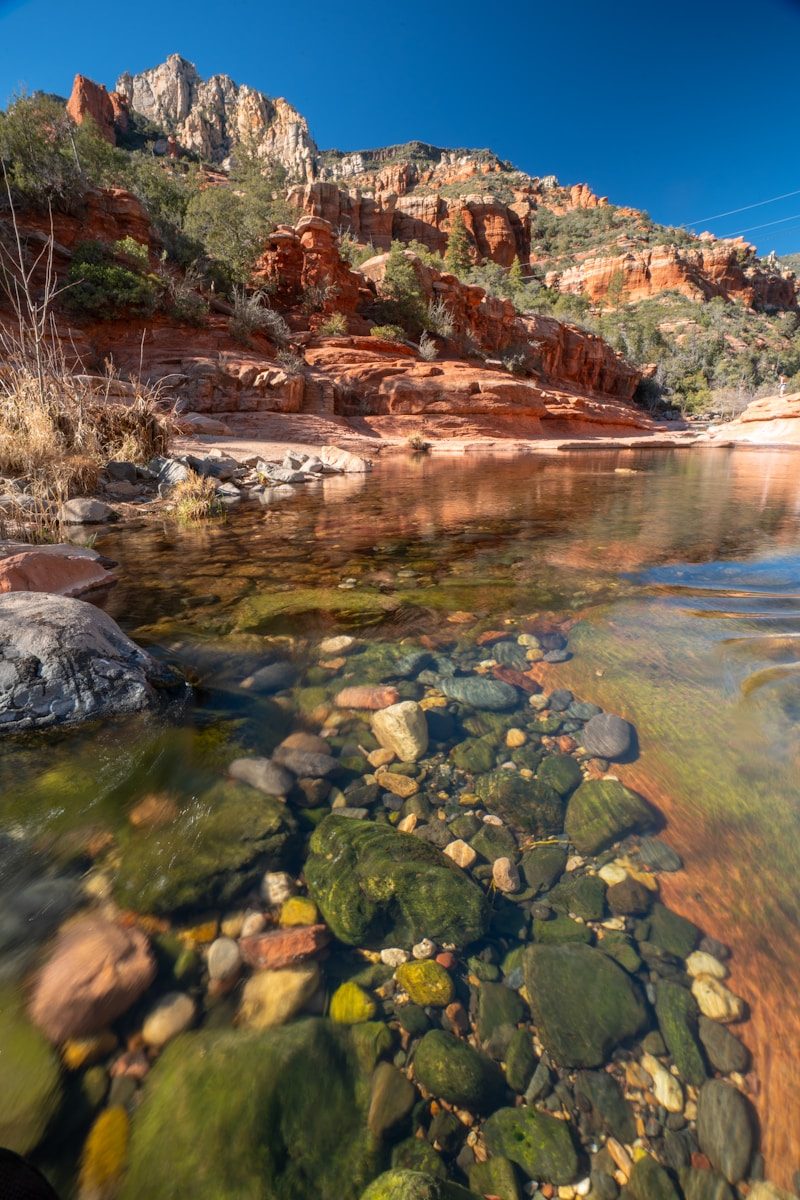 Red rock formations above a clear stream with colorful rocks.